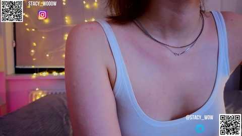 Media: A video of a woman in a white tank top, wearing a necklace, with a soft-focus background featuring fairy lights and a window.