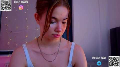 Media: Video of a young woman with light skin, brown hair tied back, wearing a white tank top and silver necklace, looking down. Background features a window with a starry night scene.