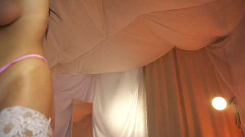 Media: A video of a woman with light skin, wearing a pink lace bra and a white veil, stands against a backdrop of beige curtains and a hanging light bulb, creating a romantic, intimate atmosphere.