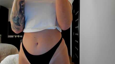 Media: Video of a light-skinned woman in black panties, lifting a white crop top to reveal her toned stomach. She has a tattoo on her left arm. Background includes a bed with a quilt, a dark wardrobe, and a partially visible nightstand.