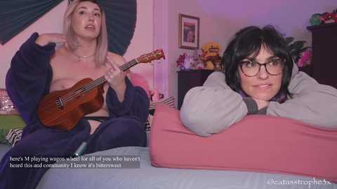 Media: A video features a blonde woman topless, playing a ukulele in a bedroom, while a woman with glasses and short black hair rests her head on a pink pillow, looking displeased.