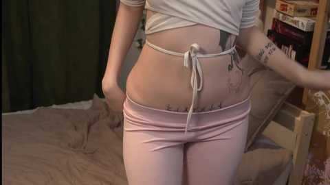 Media: Video of a young woman with fair skin, wearing a white cropped top and light pink shorts, standing in a bedroom with a messy bed and shelves in the background.