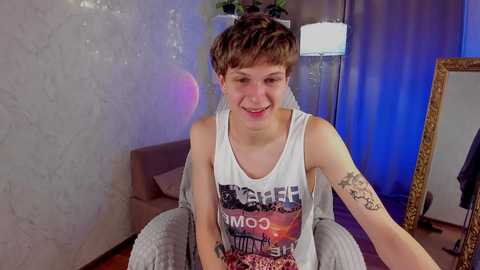 Media: A smiling, short-haired transgender woman with tattoos, wearing a white tank top with a graphic print, sits on a grey chair in a dimly lit room with a mirror and potted plant.