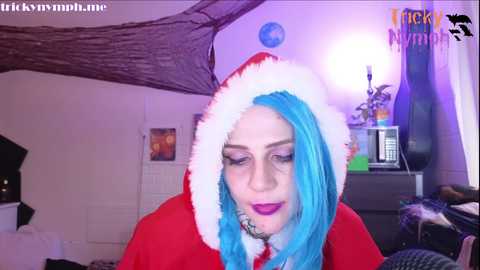 Media: Video of a woman with vibrant blue hair, wearing a red Santa hat and cape, in a colorful, slightly messy bedroom with purple walls.