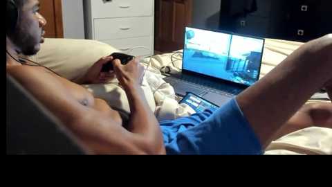 Media: Video of a shirtless, muscular Black man in blue shorts, playing video games on a laptop, with a TV showing the same game in the background, on a bed with white sheets and wooden furniture.