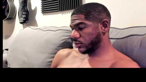 Media: Video of a shirtless, dark-skinned man with a trimmed beard and close-cropped hair, sitting on a gray couch. He appears thoughtful, with a cigarette in his mouth. The background features a vent and a black wall mount.