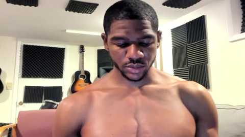 Media: Video of shirtless, muscular Black man with short hair and beard, standing in a modern, well-lit room with black acoustic guitar, amplifier, and soundproof panels on white walls.