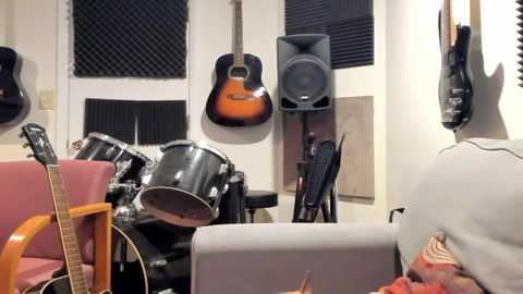 Media: A video of a cluttered music room with a pink couch, a black drum set, an electric guitar, and acoustic guitars hanging on the wall, surrounded by various musical instruments and soundproofing panels.