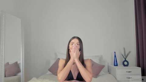 Media: Video of a young woman with long brown hair, wearing a black top, covering her face with her hands, sitting on a bed with white bedding and pillows, in a modern, minimalist bedroom with white walls, a white nightstand, and a blue vase.