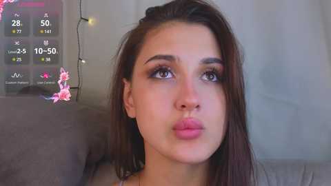 Media: Video of a young woman with fair skin, straight brown hair, and full lips, making a pouty expression. Background includes a weather display and a string of fairy lights.