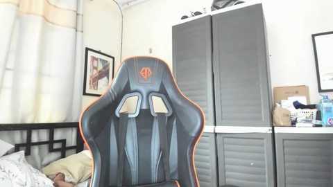 Media: Video of a dark blue gaming chair with orange accents, placed in a cluttered bedroom with beige curtains, a bed, a gray wardrobe, and various items on the floor and desk.