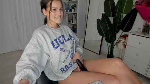 Media: Video of a smiling young woman with light skin, dark hair, wearing a gray UCLA sweatshirt, sitting on a chair, with a large plant and white furniture in the background.