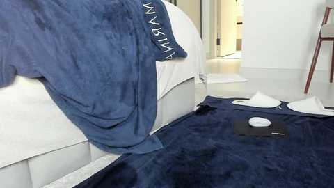 Media: Video of a person in a navy-blue bathrobe with white text on the sleeve, standing by a white bed with a dark blue blanket and a white mouse. Background shows a modern white room with a wooden chair and a partially open door.