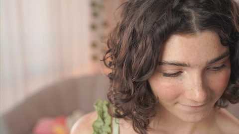 Media: Video of a young woman with curly, shoulder-length brown hair, wearing a green sleeveless top, smiling softly, set against a blurred, pastel-colored background.