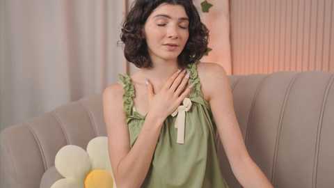 Media: Video of a young woman with curly black hair, wearing a green, ruffled tank top, hands over her heart, sitting on a beige sofa with pastel balloons.