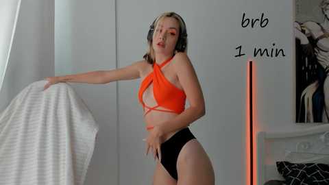 Media: Video of a young, light-skinned woman with short blonde hair, wearing a vibrant orange and black halter bikini top and high-waisted black bottoms, standing in a modern, minimalist bedroom.