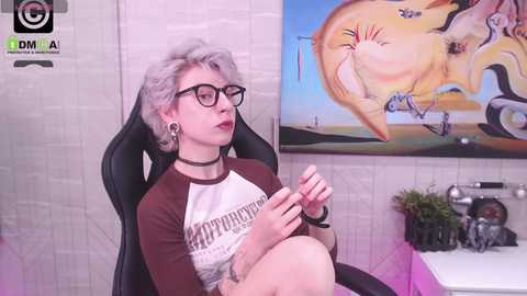 Media: A video of a pale-skinned woman with short, silver hair, glasses, and a black choker, sitting in a gaming chair, wearing a \"Hot Topic\" shirt, against a tiled wall with a colorful painting.