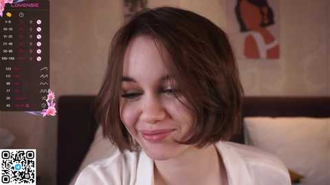 Media: Video of a young woman with short, straight brown hair, fair skin, and a slight smile, wearing a white blouse. Background includes a dark headboard, a calendar with pink flowers, and a colorful abstract painting.