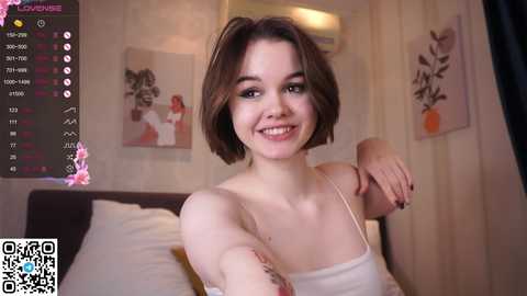 Media: Video of a smiling, fair-skinned young woman with short brown hair, wearing a white tank top, in a cozy bedroom with floral wall art.