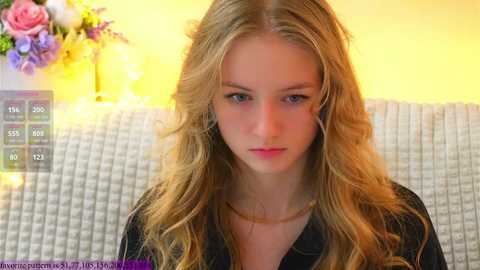 Media: A video of a young blonde woman with wavy hair, fair skin, and blue eyes, wearing a black jacket, seated indoors with warm lighting, a floral arrangement, and a white textured couch in the background.