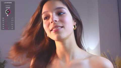 Media: A video of a young, light-skinned woman with long, wavy brown hair, wearing gold hoop earrings, smiling. The background features a blurred indoor setting with a digital calendar overlay.