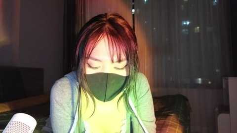 Media: Video of a woman with long red hair and a green mask, sitting in a dimly lit room, wearing a gray sweater, with a white speaker on the left.