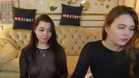 Media: Video of two young women, one with a cross necklace, wearing a sheer black top, and the other in a black sweater, seated in a beige tufted couch, with decorative pillows and wall art in the background.