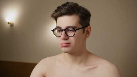 Media: Video of a young, fair-skinned man with short, dark brown hair, wearing black-framed glasses and no shirt, in a beige room with a wall-mounted light fixture.