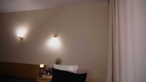 Media: Video of a simple, dimly lit bedroom with beige walls, two wall-mounted sconces, a wooden nightstand with a lamp and a potted plant, a white chair, and a black pillow.