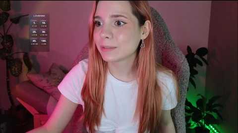 Media: A video of a young Caucasian woman with long, straight, reddish-brown hair, wearing a white T-shirt, sitting indoors, lit with a green and pink light, and looking slightly to the right.