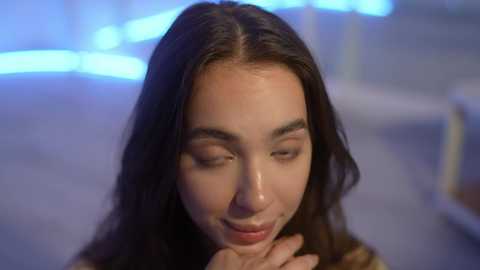 Media: A video of a young woman with long, dark brown hair, fair skin, and closed eyes, softly smiling, with hands gently touching her face. The background features a blurred, blue-tinted, modern indoor setting.