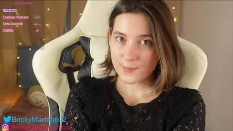 Media: A video of a young woman with fair skin, shoulder-length brown hair, and brown eyes, wearing a black lace dress, seated in a white gaming chair.