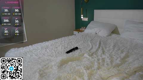 Media: Video of a modern bedroom with a white bed covered in a plush, light-colored rug. A black remote control lies on the bed. The teal wall features a mounted weather display and a gold lamp.