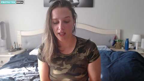 Media: Video of a young woman with wet hair, wearing a camouflage t-shirt, sitting on a bed with blue sheets, in a dimly lit, messy bedroom.