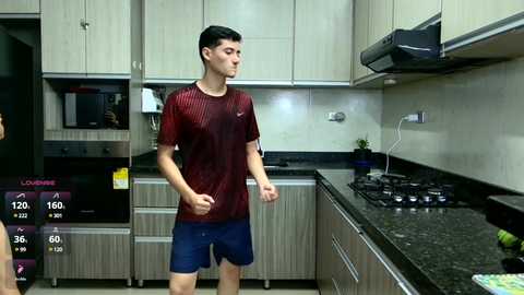 Media: Video of a young man with short dark hair in a modern kitchen, wearing a red and blue sports shirt and shorts, standing near a black countertop, with a digital screen showing workout stats.