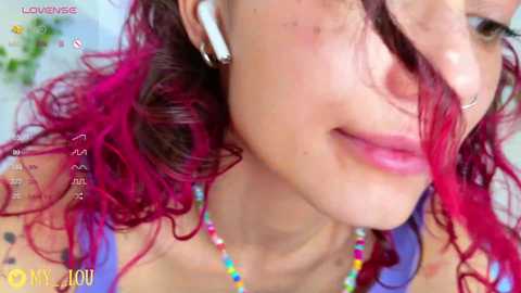 Media: Close-up video of a young woman with light skin, pink lips, and vibrant pink and purple hair, wearing a nose ring and colorful beaded necklace.