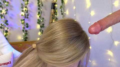 Media: Video of a blonde-haired woman's head in profile, with a hand touching her hair, against a background of fairy lights and green ivy, creating a cozy, intimate atmosphere.