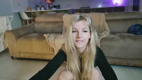 Media: Video of a smiling, fair-skinned, blonde woman with long hair, wearing a black top, sitting on a beige sofa in a dimly lit living room.