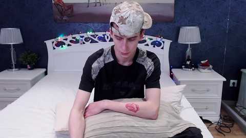 Media: Video of a young man with fair skin and a bandaged wrist, wearing a camo cap and black T-shirt, lying on a bed in a dimly lit bedroom with white furniture and a decorative headboard.