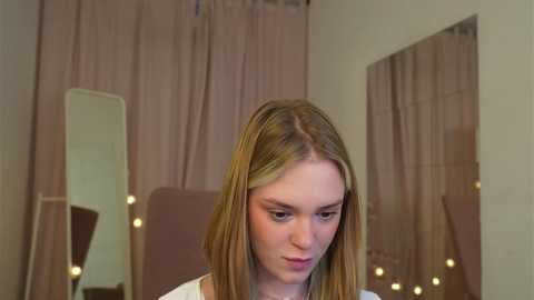 Media: Video of a young Caucasian woman with light skin and long, straight, blonde hair, wearing a white top, standing in front of a mirror with a white background and pink curtains.