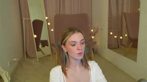 Media: Video of a young Caucasian woman with straight, shoulder-length blonde hair, wearing a white top, standing in a softly lit, vintage-style dressing room with pink curtains, mirrors, and a chair.