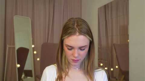 Media: Video of a young Caucasian woman with straight, light brown hair, wearing a white top, sitting in front of a mirror with pink curtains and round light bulbs.