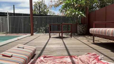 Media: Video of a sunny backyard with a wooden deck, striped cushions, and a red metal chair near a potted plant. A fenced-in pool is visible on the left, and lush greenery surrounds the area.