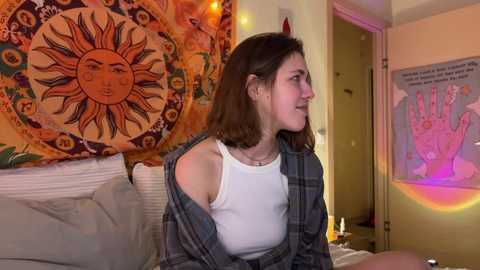 Video of a young woman with shoulder-length brown hair, wearing a white tank top and a plaid shirt, seated in a cozy room with colorful tapestries and warm lighting.