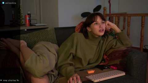 Media: Video of a young woman with light skin, dark hair, wearing a green turtleneck and beige pants, lying on a couch, looking contemplative.