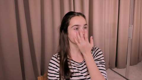 Media: Video of a young girl with long dark hair, wearing a striped shirt, covering her face with her hand, in a room with beige curtains and a cream carpet.