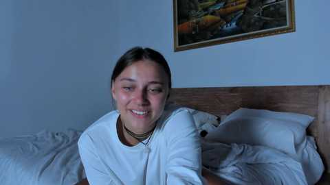 Media: Video of a young woman with light skin and dark hair, smiling, in a white shirt, lying on a bed with rumpled white sheets. A colorful painting of fish is on the wall behind her.