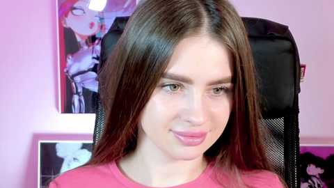 Media: Video of a young woman with straight, shoulder-length brown hair, fair skin, and a pink shirt, sitting in a black gaming chair. The background features anime posters with bright colors and a pink wall.