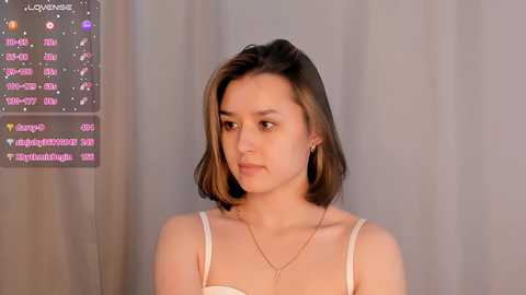 Media: A video of a young woman with medium-length brown hair, wearing a white spaghetti-strap top and a delicate gold necklace. She stands against a plain gray curtain, with a digital screen displaying social media notifications in the background.