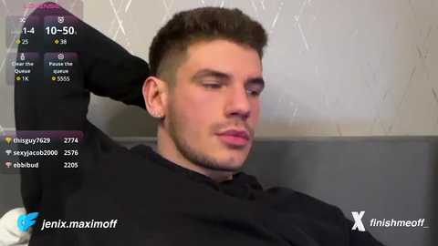 Media: Video of a young man with short brown hair, fair skin, and a clean-shaven face, wearing a black shirt, sitting on a grey couch, with a \"X\" watermark and social media handles in the corner.